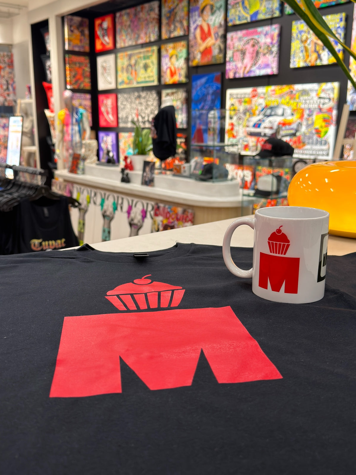 A branded t-shirt and matching mug printed with a bakery logo, on the counter at Urban Customz